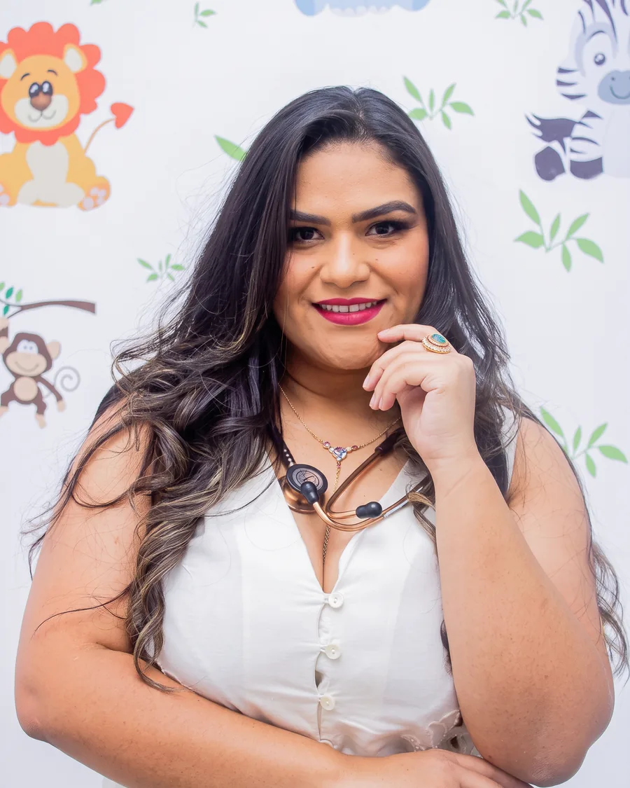 Dra. Bruna Rafaela Carvalho no consultório, diante da parede ilustrada com bichos, com o estetoscópio no pescoço.