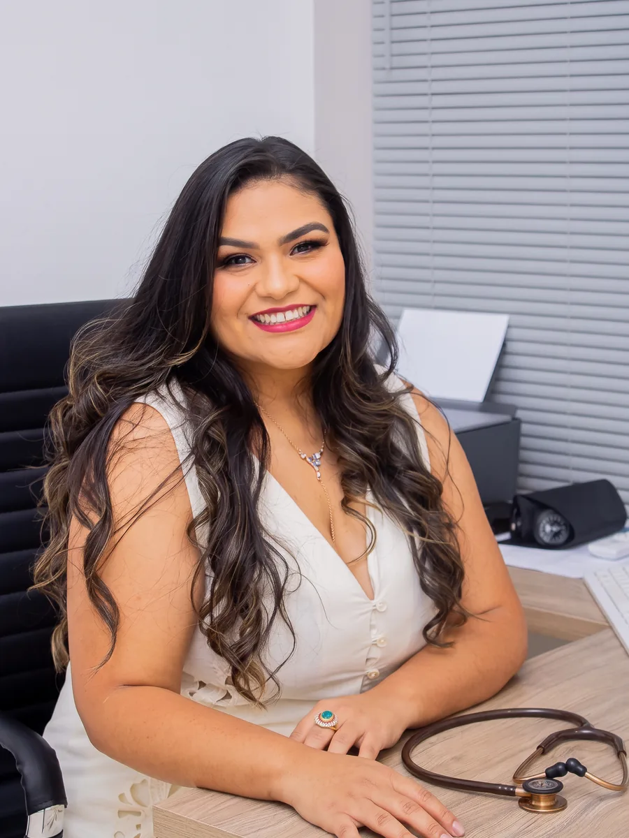 Dra. Bruna Rafaela Carvalho sentada à mesa do consultório, com o estetoscópio ao lado.